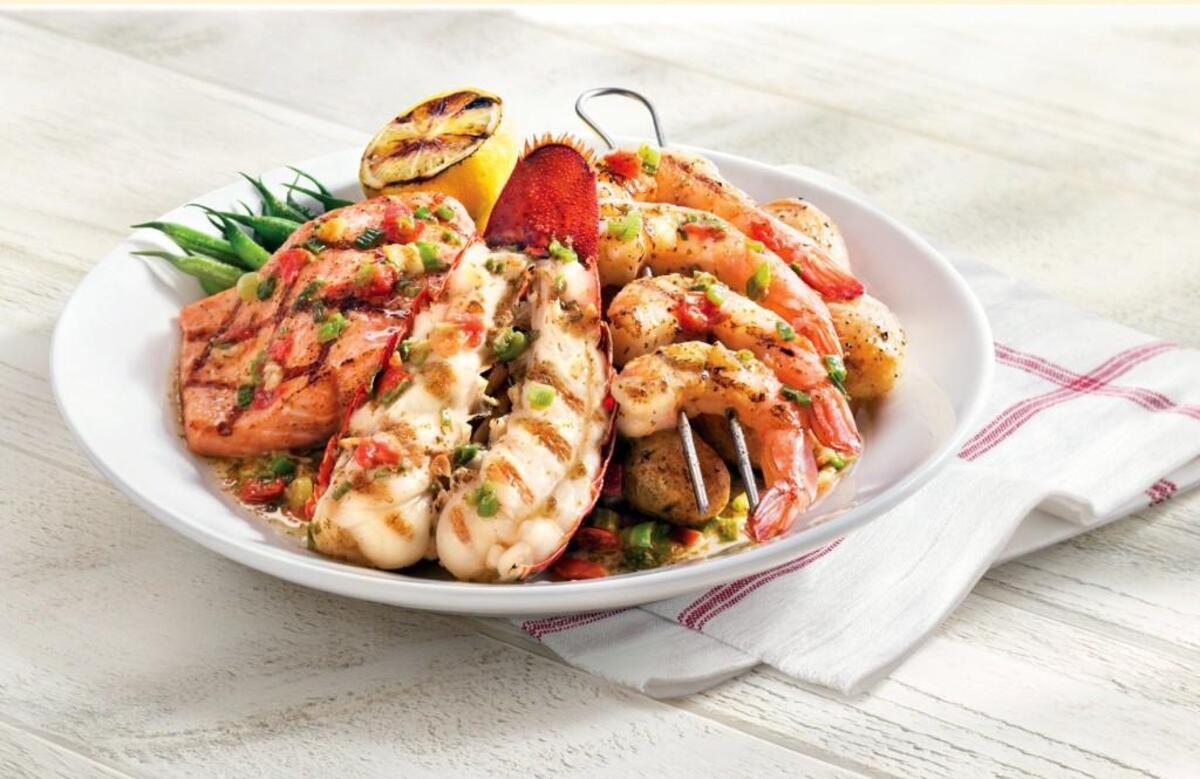 A large white platter on a white wooden table holds a meal of grilled salmon, a lobster tail cut in half, and a skewer of shrimp. The seafood is drizzled with a sauce and accompanied by green beans and a grilled lemon half. A white napkin with red stripes is tucked under the plate.