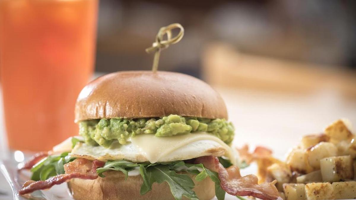 Elevated Egg Sandwich with an over easy egg, bacon, Gruyere cheese, fresh smashed avocado, lemon crema and arugula on a brioche bun, served with fresh, seasoned potatoes.