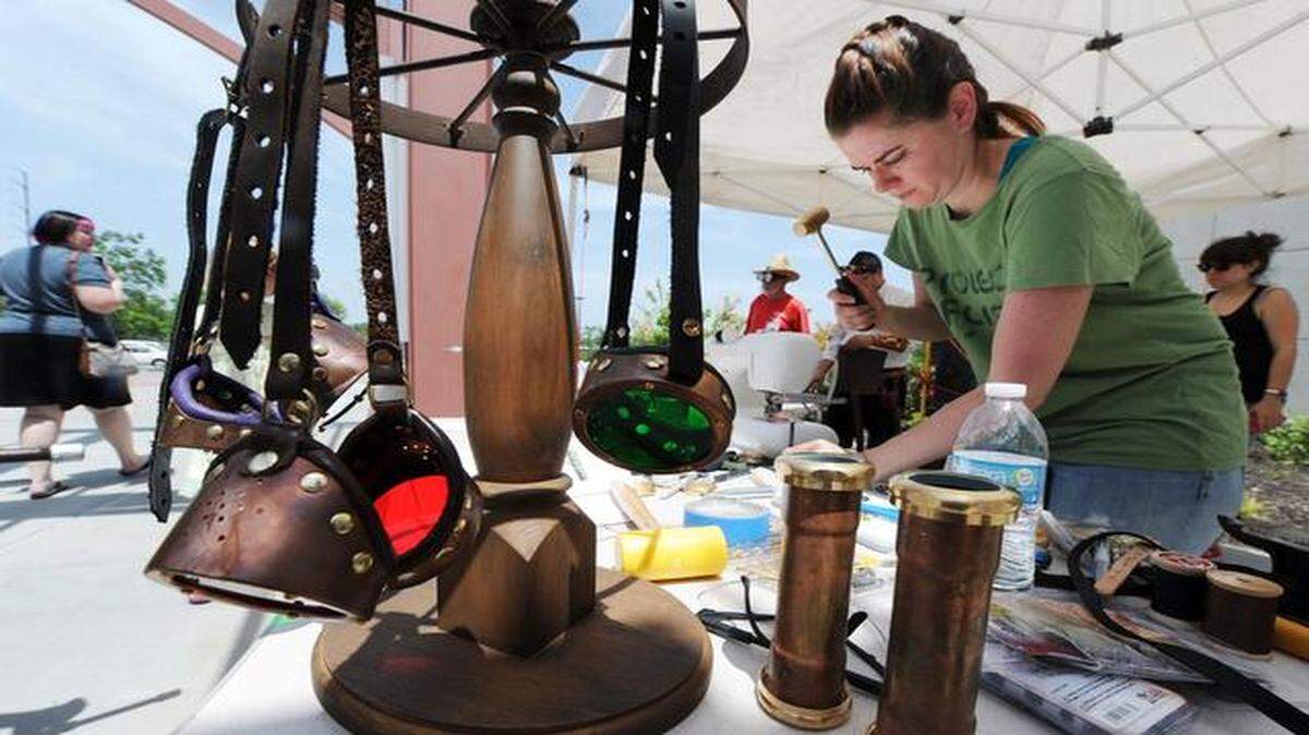 
Kat Kaufman, right, of Parsons, Kan., hammers a brass fitting into place while making a pair of steampunk goggles, like the ones hanging on the display at left, during the Johnson County Public Library’s Second Saturday event on July 12.
