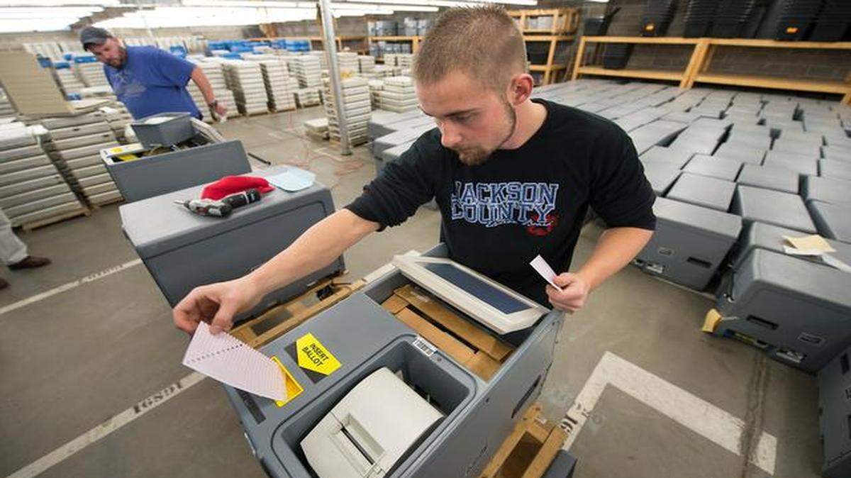 
Dakota Bossow, (center), an election technician, worked on an Inka Vote precinct ballot-counting machine and prepared to run a test ballot through the machine last week at the Jackson County Election Board’s warehouse in Independence. All 325 of the ballot counters, as well as the voting machines (stacked in background) are maintained and stored at the facility.
