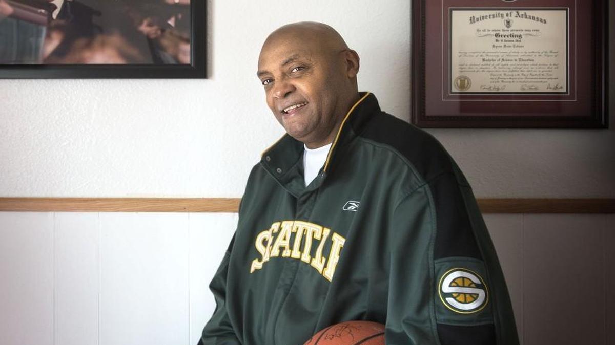 Former Seattle SuperSonics player Dean Tolson wore his old warmup jacket in his Tacoma, Wash., home in front of a wall that had a photo of him graduating from the University of Arkansas and his two diplomas from the school.
